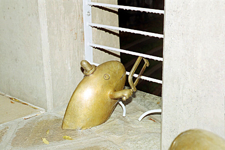 [A brass fish with wearing a little hat is part-way out of the cement using a saw with its little arm on some wire fencing.]